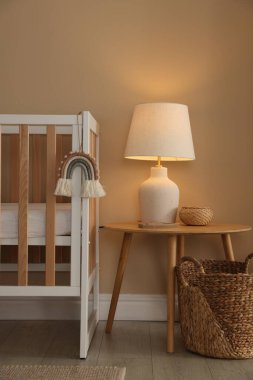 İçerideki bej duvarın yanında beşik, masa, lamba ve dekor. Şık bir bebek odası.