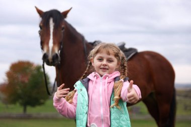 Equine assisted therapy. Little girl showing thumbs up and beautiful horse outdoors, selective focus. Lovely domesticated pet
