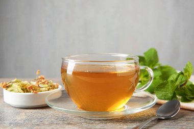 Cam fincanda lezzetli çay, ıhlamur, nane ve çay kaşığı ahşap masada gri arka planda, yakın planda.