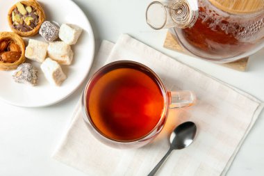Cam fincanda lezzetli çay, çay kaşığı, Türk tatlısı ve çaydanlığı beyaz masada, düz yerde.