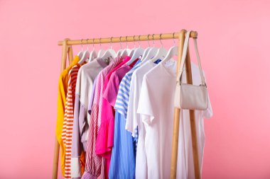 Farklı kıyafetler ve çantalar pembe arka planda asılı, yakın plan.