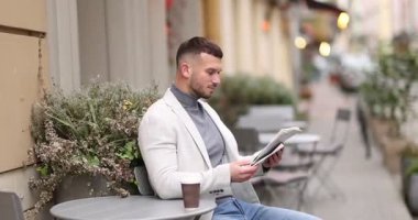 Genç adam açık kafede kahve içerken gazete okuyor.