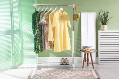 Farklı kadın kıyafetleri askıda, aynada, yan masada ve yeşil duvarın yanında asılı duruyor.