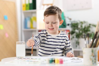 Gülümseyen küçük çocuk fırçayla beyaz masaya çizim yapıyor.