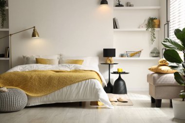 Yatak odasında çarşaflı, komodinli, puf ve raf üniteli büyük bir yatak.