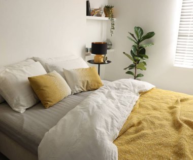 Yatak odasındaki beyaz duvarın yanında çarşaflı, komodinli büyük bir yatak.