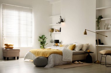 Yatak odasındaki beyaz duvarın yanında çarşaflı, komodinli, puflu ve lambalı büyük bir yatak.