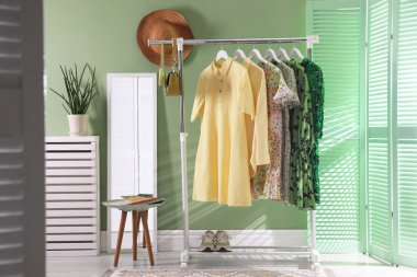 Farklı kadın kıyafetleri askıda, aynada, yan masada ve yeşil duvarın yanında asılı duruyor.