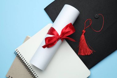 Diploma, mezuniyet şapkası ve açık mavi arkaplan üzerine not defterleri.