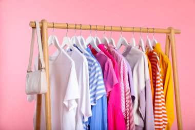 Farklı kıyafetler ve çantalar pembe arka planda asılı, yakın plan.