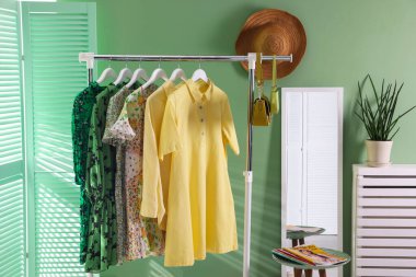 Farklı kadın kıyafetleri askıda, aynada, yan masada ve yeşil duvarın yanında asılı duruyor.