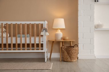 İçerideki bej duvarın yanında beşik, masa, lamba ve dekor. Şık bir bebek odası.