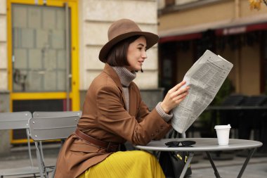 Şapkalı ve takım elbiseli gülümseyen kadın açık kafede gazete okuyor.