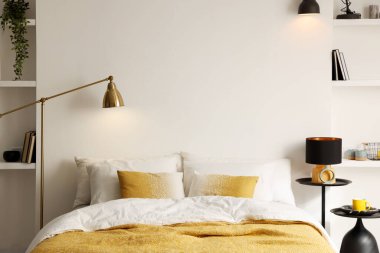 Yatak odasındaki beyaz duvarın yanında çarşafları, komodinleri ve lambaları olan büyük bir yatak.