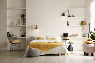 Yatak odasında çarşaflı, komodinli, puf ve raf üniteli büyük bir yatak.