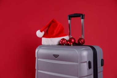 Noel tatili. Kırmızı arka planda Noel Baba şapkası ve takıları olan gri bavul, yakın plan. Metin için boşluk