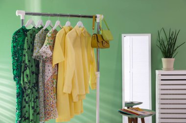 Farklı kadın kıyafetleri askıda, aynada, yan masada ve yeşil duvarın yanında asılı duruyor.