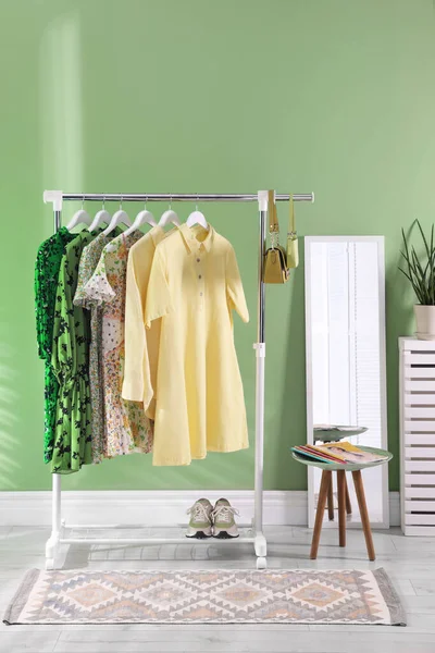 Farklı kadın kıyafetleri askıda asılı, ayna ve yan masa yeşil duvarın yanında.