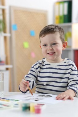Gülümseyen küçük çocuk fırçayla beyaz masaya çizim yapıyor.