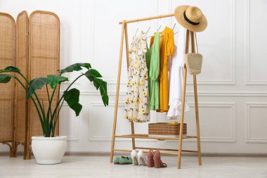 Farklı kadın kıyafetleri askıda asılı, katlanan ekran ve beyaz duvarın yanında bitki örtüsü