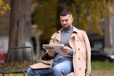 Genç adam dışarıda sandalyede gazete okuyor.