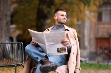 Genç adam dışarıda sandalyede gazete okuyor.