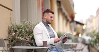 Genç adam açık kafede kahve içerken gazete okuyor.