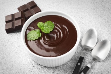 Erimiş çikolata kasede nane, parçalar ve kaşıklar açık gri masada, yakın plan.