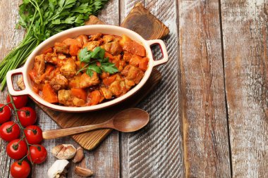 Pişirme kabında nefis pişirilmiş güveç, taze malzemeler ve tahta masada kaşık, düz yatak. Metin için boşluk