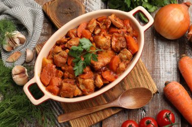 Pişirme kabında nefis pişirilmiş güveç, taze malzemeler ve tahta masada kaşık, düz uzanmış.