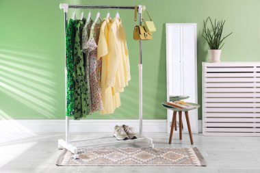 Farklı kadın kıyafetleri askıda, aynada, yan masada ve yeşil duvarın yanında asılı duruyor.