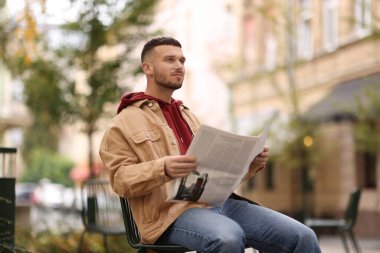 Genç adam dışarıda sandalyede gazete okuyor.