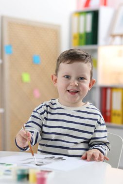 Gülümseyen küçük çocuk fırçayla beyaz masaya çizim yapıyor.