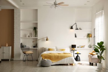 Yatak odasında çarşaflı, komodinli, puf ve raf üniteli büyük bir yatak.