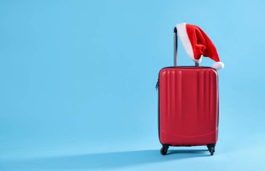 Noel tatili. Açık mavi zemin üzerinde Noel Baba şapkası olan kırmızı bavul, metin için boşluk