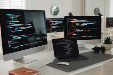 Ofisteki beyaz masada çift monitörlü, dizüstü bilgisayarı ve kulaklıklı programcının işyeri