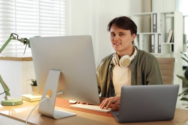 Ofisteki ahşap masada bilgisayarla çalışan kulaklıklı gülen programcı.