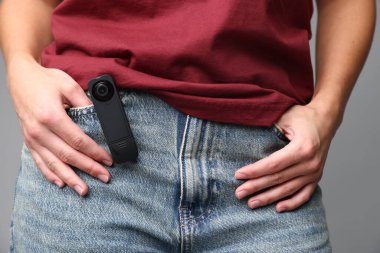 Cebinde gizli kamera olan genç bir kadın açık gri arka planda, yakın plan.