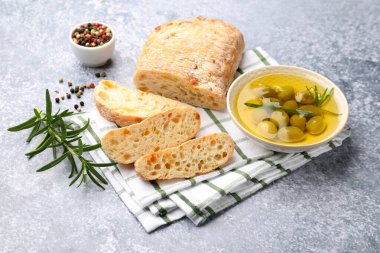 Taze kesilmiş ciabatta, yağ, zeytin, biberiye ve biberiyeler gri desenli masada, yakın plan.