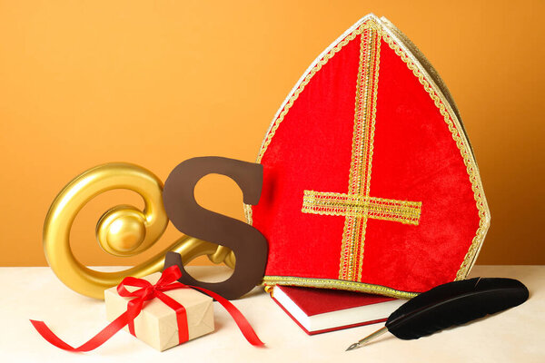 День святого Николая (Sinterklaas). Шоколадная буква S, mitre, crosier, подарок, блокнот и перо на белом столе