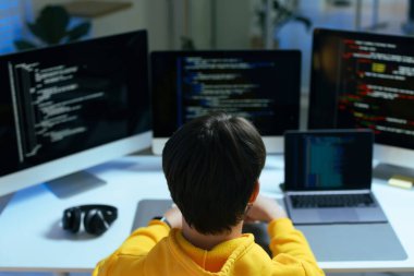 Ofisteki beyaz masada üç monitörlü ve laptoplu bilgisayar üzerinde çalışan programcı.