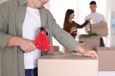 Taşınma günü. Ofiste farklı eşyalar paketleyen bir grup işçi, yapıştırıcı bant ile karton kutuları bantlayan adama odaklan.