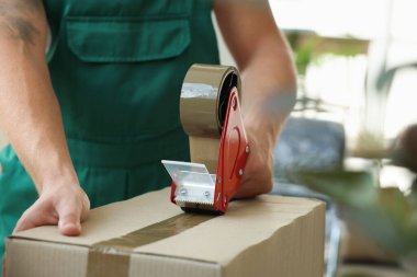 Taşınma günü. Genç adam, ofiste yapıştırıcı bant kutusuyla karton kutuyu bantlıyor.