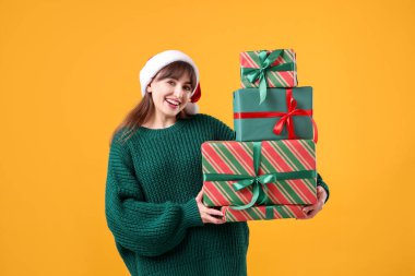 Sarı arka planda bir sürü hediye kutusu olan, Noel Baba şapkalı gülümseyen kadın. Noel ve Yeni Yıl Kutlaması