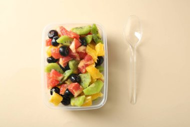 Plastik kabın içinde lezzetli meyve salatası ve bej arka planda kaşık, düz yatak. Sağlıklı çerez