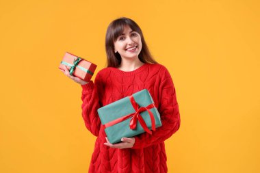 Sarı arka planda hediye kutularıyla kırmızı kazaklı, gülümseyen kadın. Noel ve Yeni Yıl Kutlaması