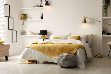 Yatak odasında çarşaflı, komodinli, puf ve raf üniteli büyük bir yatak.