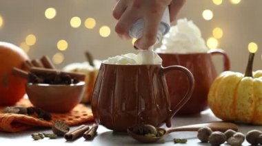 Açık gri masada, bulanık ışıklara karşı lezzetli balkabaklı latteye krema ekleyen kadın, yakın plan. Bokeh etkisi