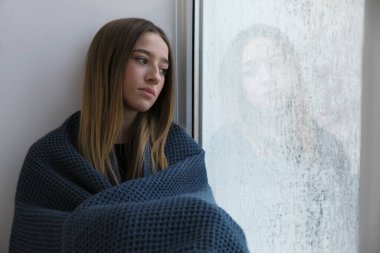 Depresyondaki genç kız evinin penceresinin yanında battaniyeye sarılmış, mesaj atmak için yer var.