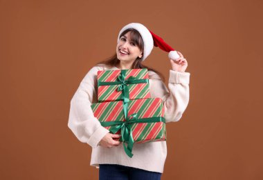 Kahverengi arka planda hediye kutularıyla Noel Baba şapkalı gülümseyen kadın. Noel ve Yeni Yıl Kutlaması